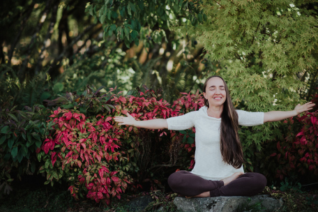 Beautiful smiling women sitting in a garden with her eyes closed and her arms open. Contact Embrace Ayurveda on beautiful community of Bainbridge island, Washington.